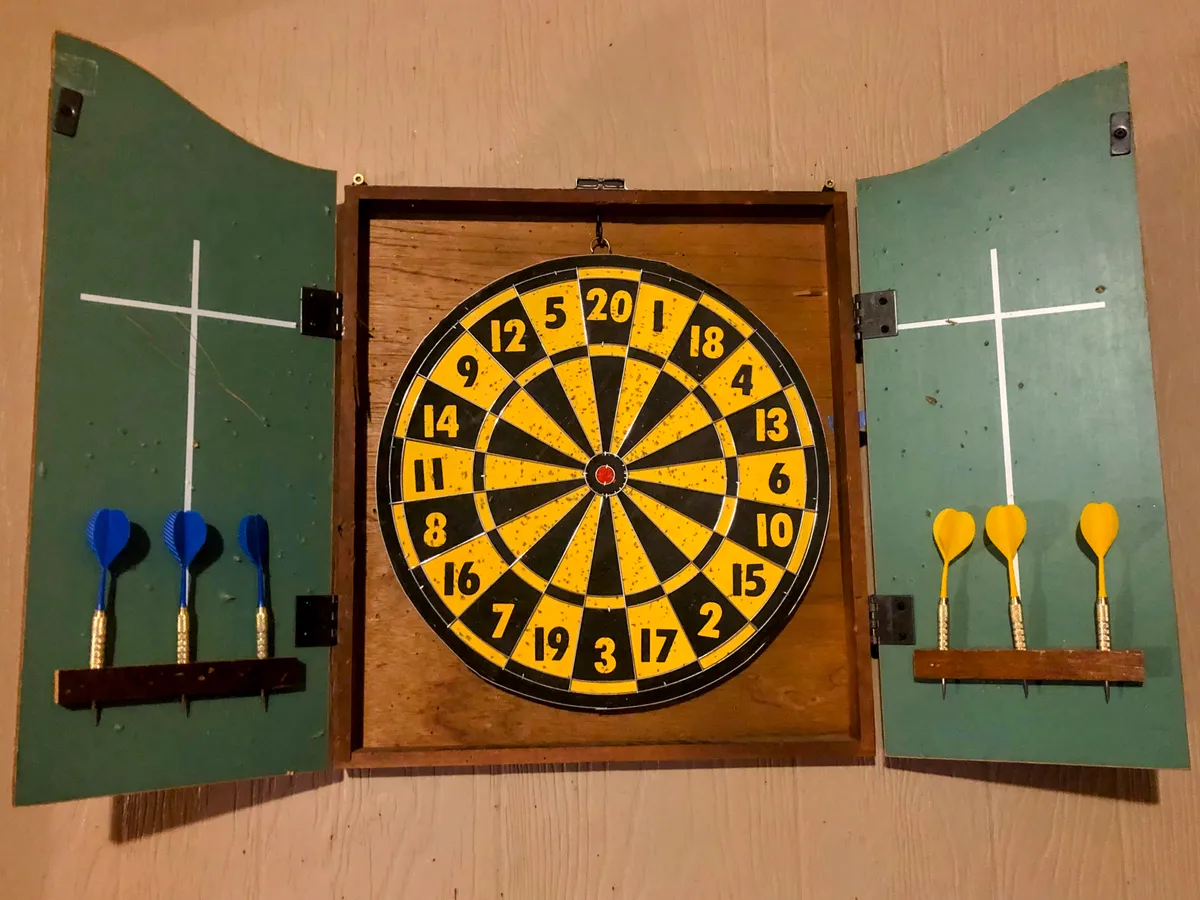 A classic dartboard mounted on a wooden cabinet awaits the next game, with darts neatly arranged and subtle wood grain textures providing a traditional game room feel.