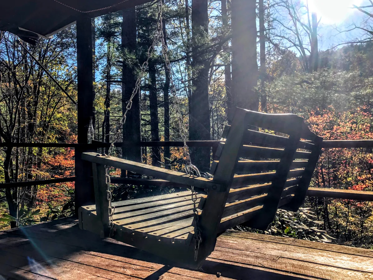 The image showcases a cozy wooden swing nestled under mature trees, surrounded by vibrant, colorful foliage. The scene exudes tranquility, inviting guests to relax, read, or simply soak in the natural beauty of the mountainside setting.