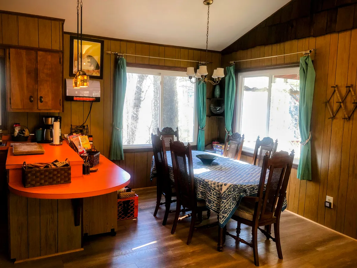 The image presents a dining area with a prominent farmhouse-style dining table, kitchen counter, large windows flooding the space with natural light, and a stylish lighting fixture overhead. The space exudes warmth and encourages family meals and social gatherings.