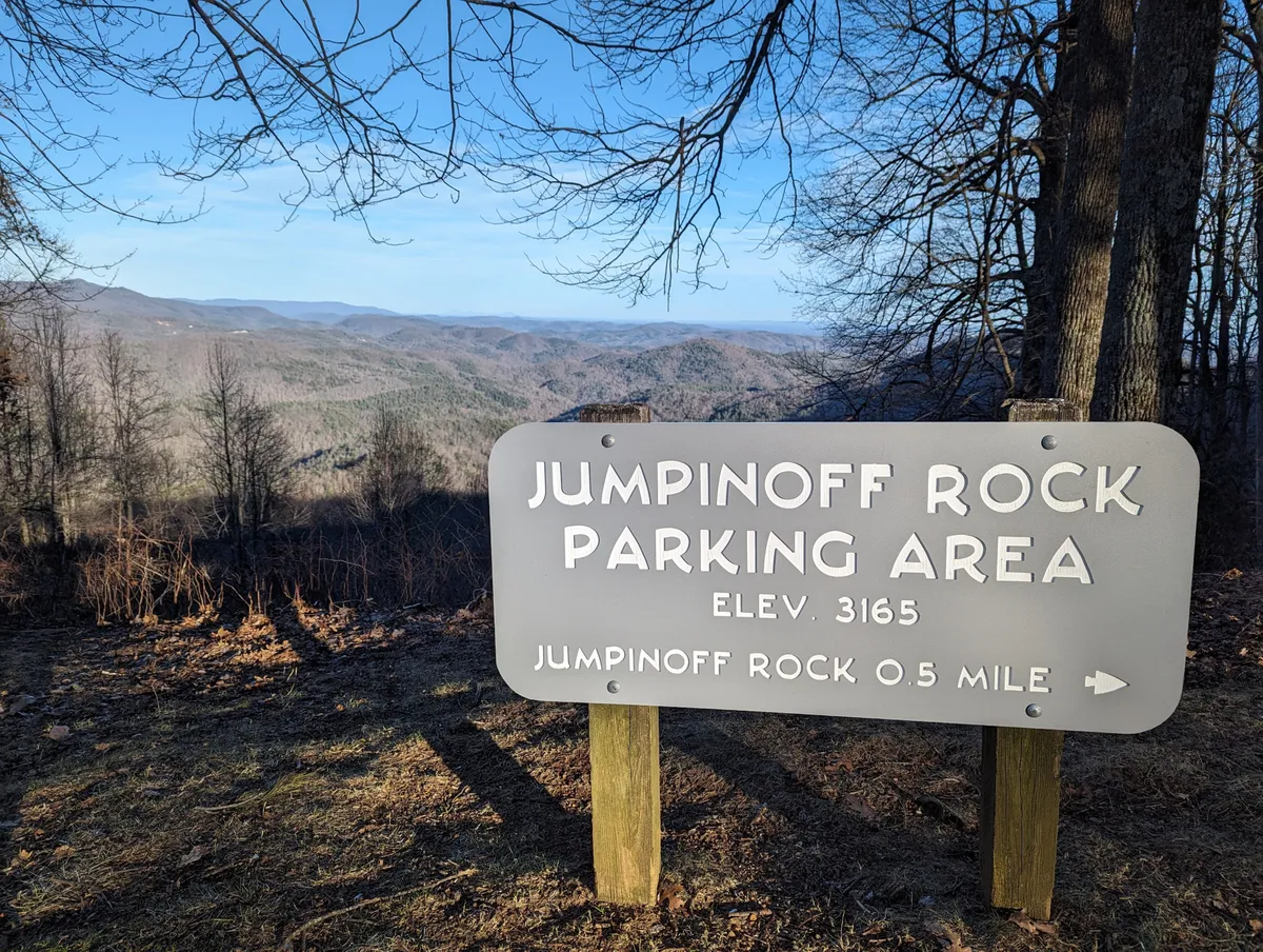The image features a sign indicating the Jumpinoff Rock Parking Area with a breathtaking mountainous landscape in the background. The scene is tranquil, with a clear view of the natural surroundings, suggesting access to popular hiking routes and scenic overlooks nearby.