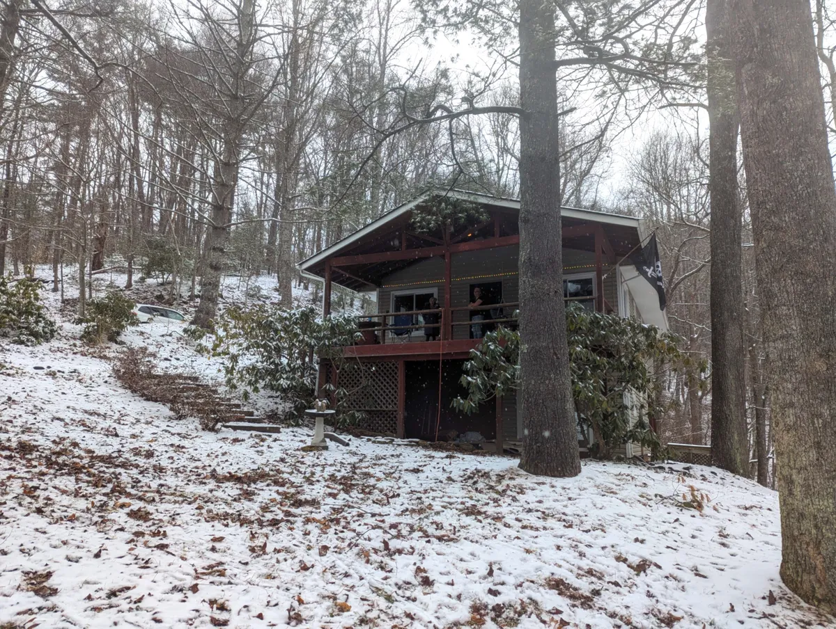 An exterior shot of the house enveloped by trees and a snow-covered yard, with hints of activity like relaxing on the porch or viewing the outdoors from inside. The tranquil, peaceful mood aligns with the idea of a cozy retreat during inclement weather.