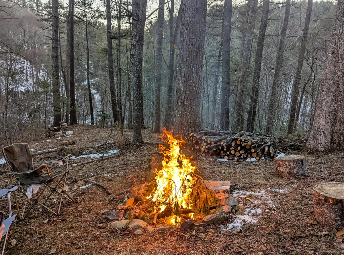 A welcoming campfire setup on a natural patch, complete with a folding chair, a stack of firewood, and tall trees bordering the space. The glow from the fire signals warmth and evening relaxation, perfect for gathering to share stories or stargaze after a busy day in town.