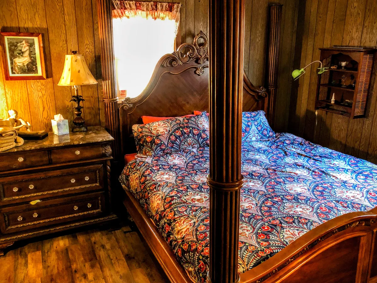 A master bedroom featuring an ornate, possibly four-poster, bed with a colorful bedspread, accompanied by a wooden dresser and a table lamp. Warm light filters into the room, highlighting the inviting and cozy atmosphere.