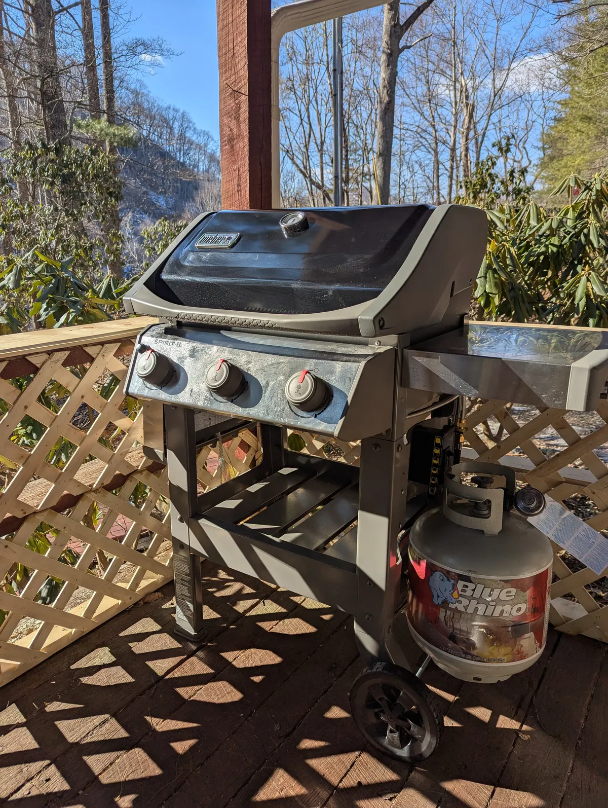 This image features a grill positioned on a wooden deck, surrounded by lush greenery. The setup hints at relaxed outdoor dining, with the grill ready for a meal and nature providing a scenic backdrop. The scene feels casual and inviting, ideal for enjoying fresh air and good food after a day out.