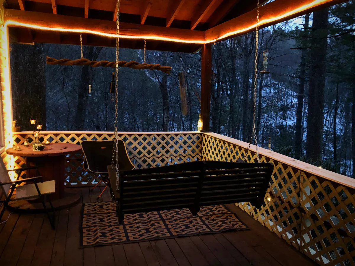 A tranquil porch scene featuring a cozy swinging bench and a wooden table, with warm string lights overhead. The ambiance suggests a relaxing spot for couples to unwind together, enjoy drinks, or gaze at the night sky while surrounded by the peace of nature.