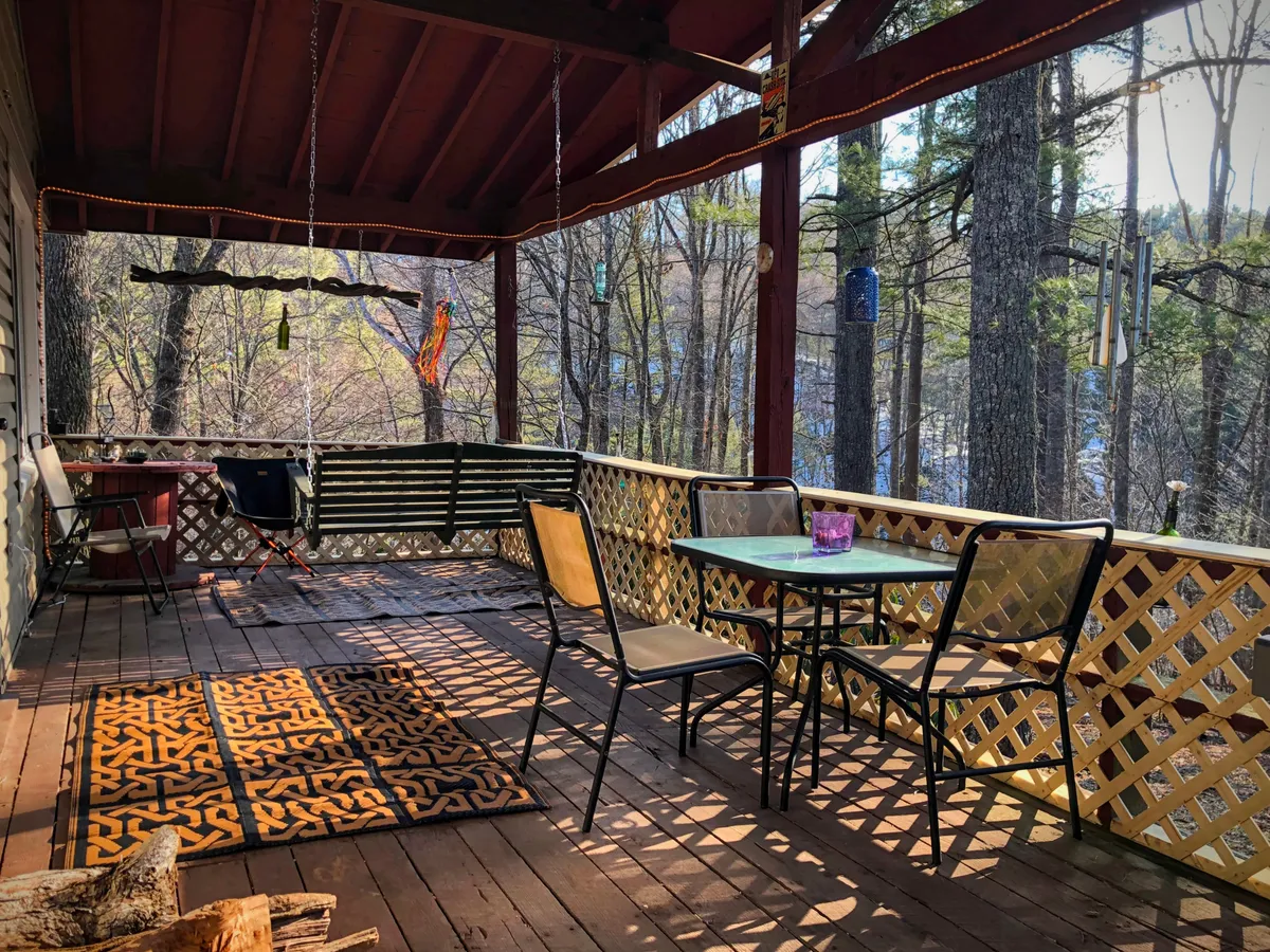 A swinging bench and an outdoor table with chairs sit atop a porch or deck, accented by a decorative rug and framed by natural surroundings. The atmosphere is serene and inviting, ideal for relaxing with a book, sharing drinks, or taking in the sunset.