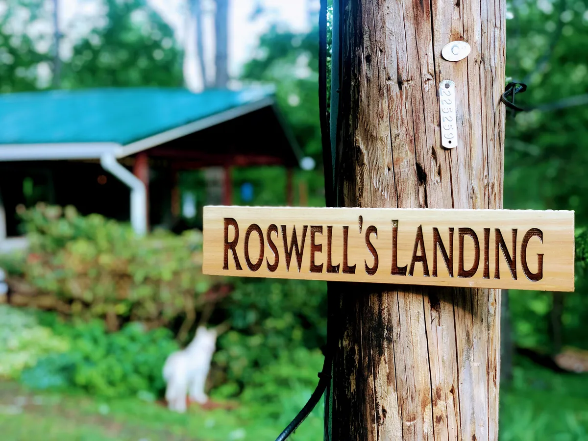 This image features a wooden sign bearing the 'Roswell's Landing' inscription, set against a backdrop of lush greenery and a glimpse of the property's exterior. The visible part of the building and surrounding natural setting create an inviting entry point for the property.