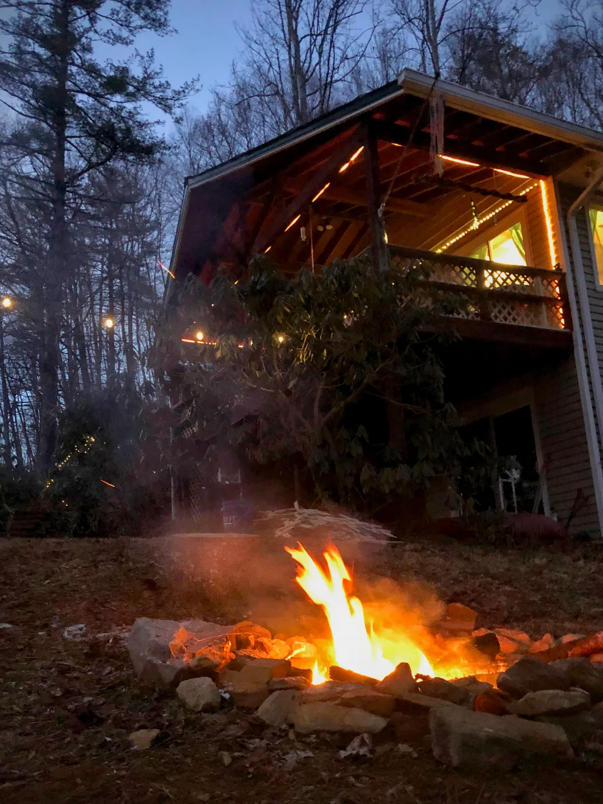 A glowing campfire burns in the yard with a house and porch lights visible in the background. Trees and a landscaped yard frame the scene, capturing a group-friendly space perfect for marshmallow roasting, storytelling, and stargazing after sunset.