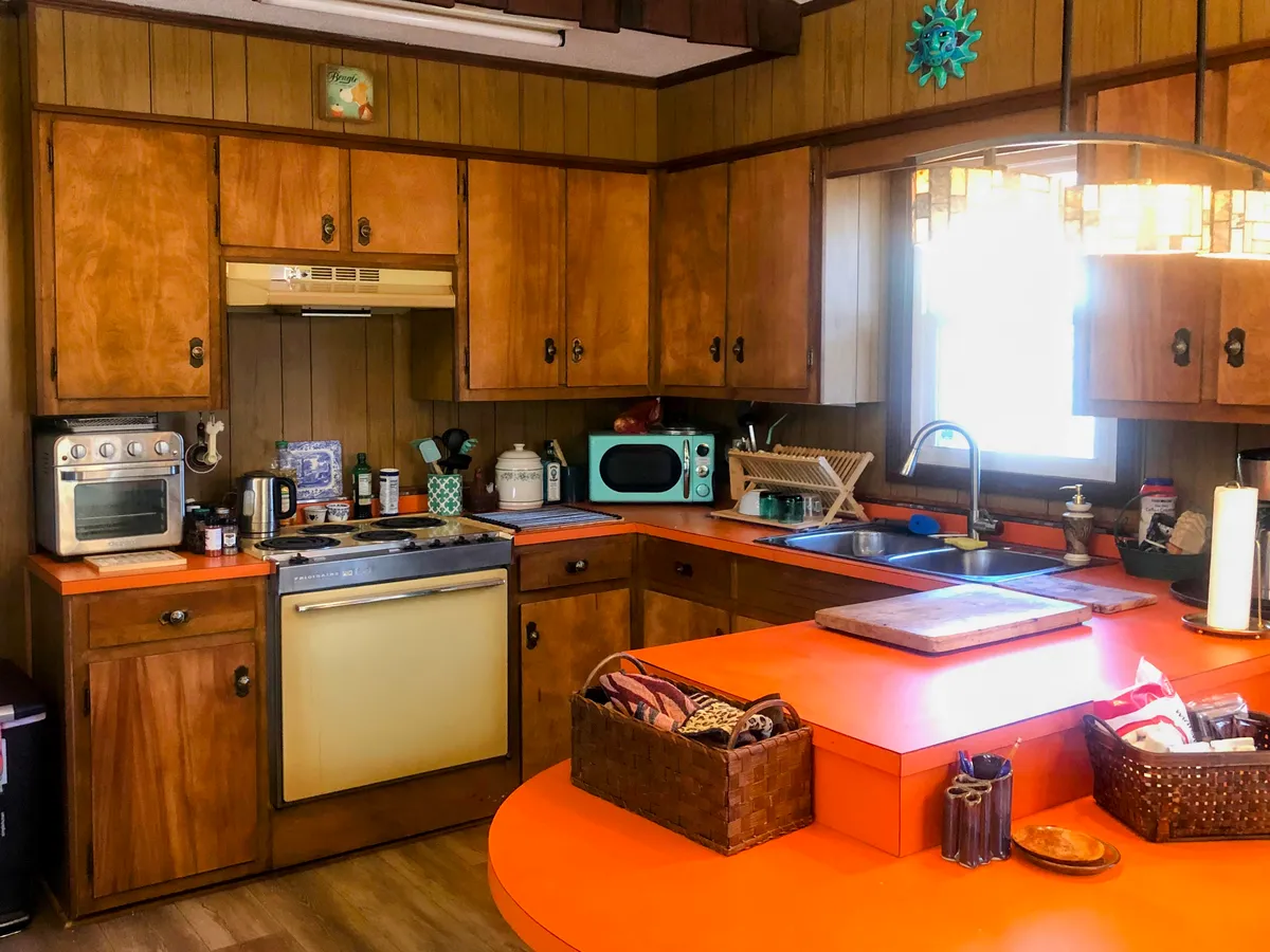 This kitchen scene features an eye-catching orange countertop, wooden cabinets, various modern kitchen appliances, and a window allowing natural light to brighten the space. The mix of retro charm and up-to-date comforts is evident in the decor and appliances.